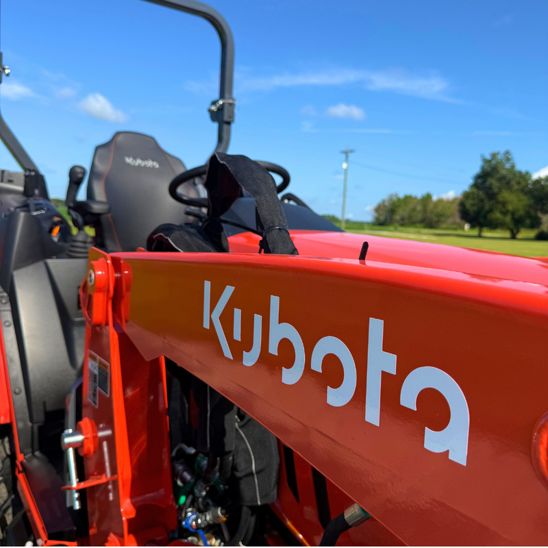 Close-up of a red Kubota tractor arm with the white Kubota logo, set against an outdoor background of blue sky and green grass.