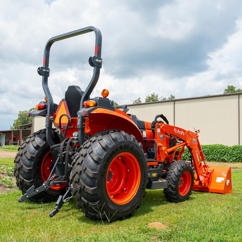 An orange Kubota compact tractor with a front loader attachment, parked on a grassy field under a cloudy sky with a building in the background.