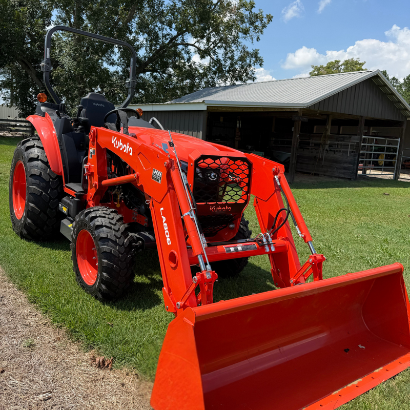 A red Kubota tractor with a front loader and bucket attachment parked on a grassy farm field, with a barn and trees in the background.